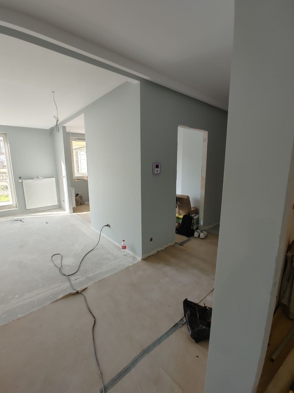 Wnętrze w trakcie prac wykończeniowych: świeżo pomalowane ściany w odcieniach szarości, widoczne kable elektryczne, zabezpieczona podłoga, butelka wody i torba z narzędziami w tle.