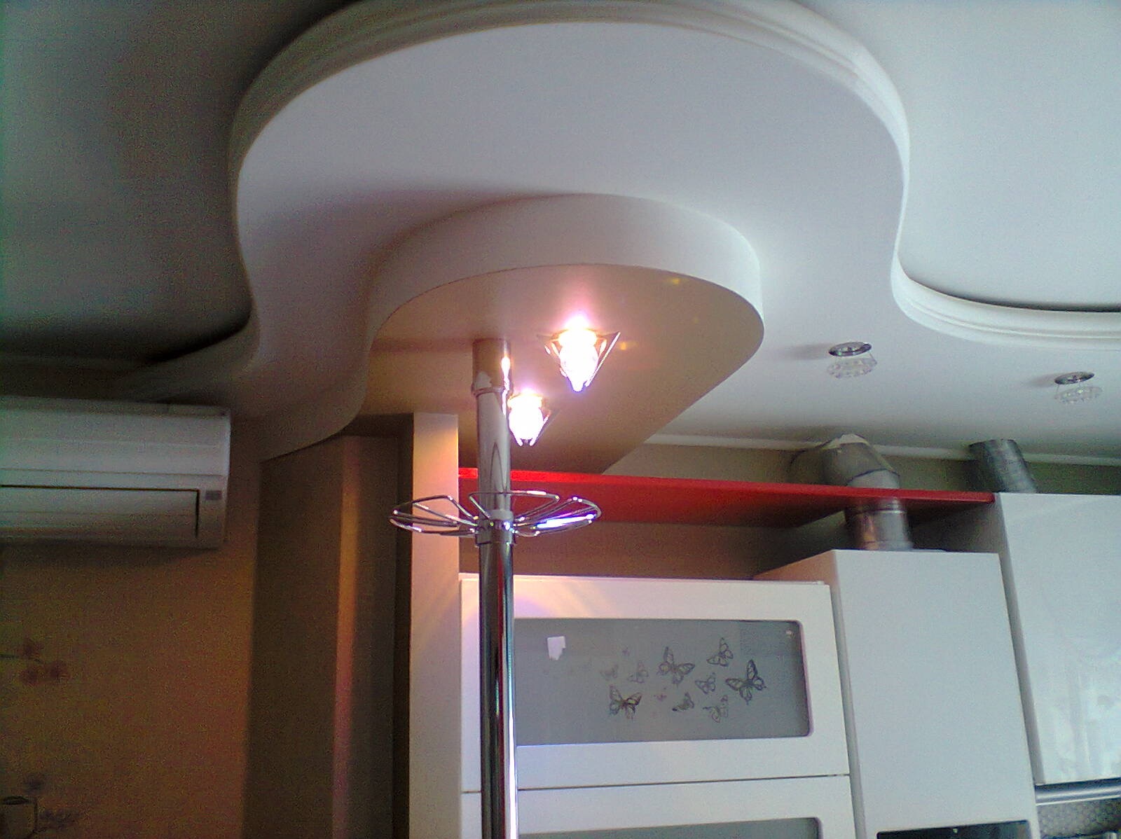 Wnętrze z dekoracyjnym sufitem podwieszanym z oświetleniem punktowym i centralnym, widoczny fragment klimatyzacji i szafki kuchennej z motywem motyli.