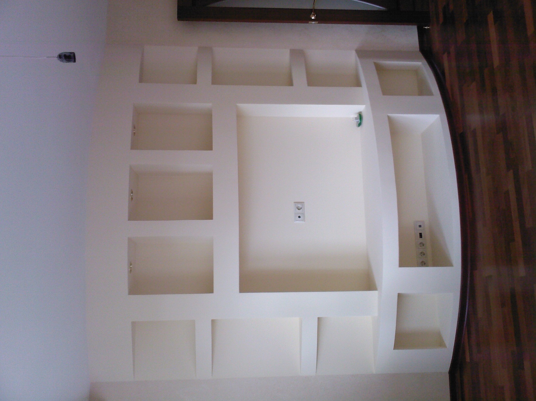 Ściana z wnękami i gładzią w odcieniach bieli, z widocznymi gniazdkami elektrycznymi i drewnianą podłogą parkietową w tle.