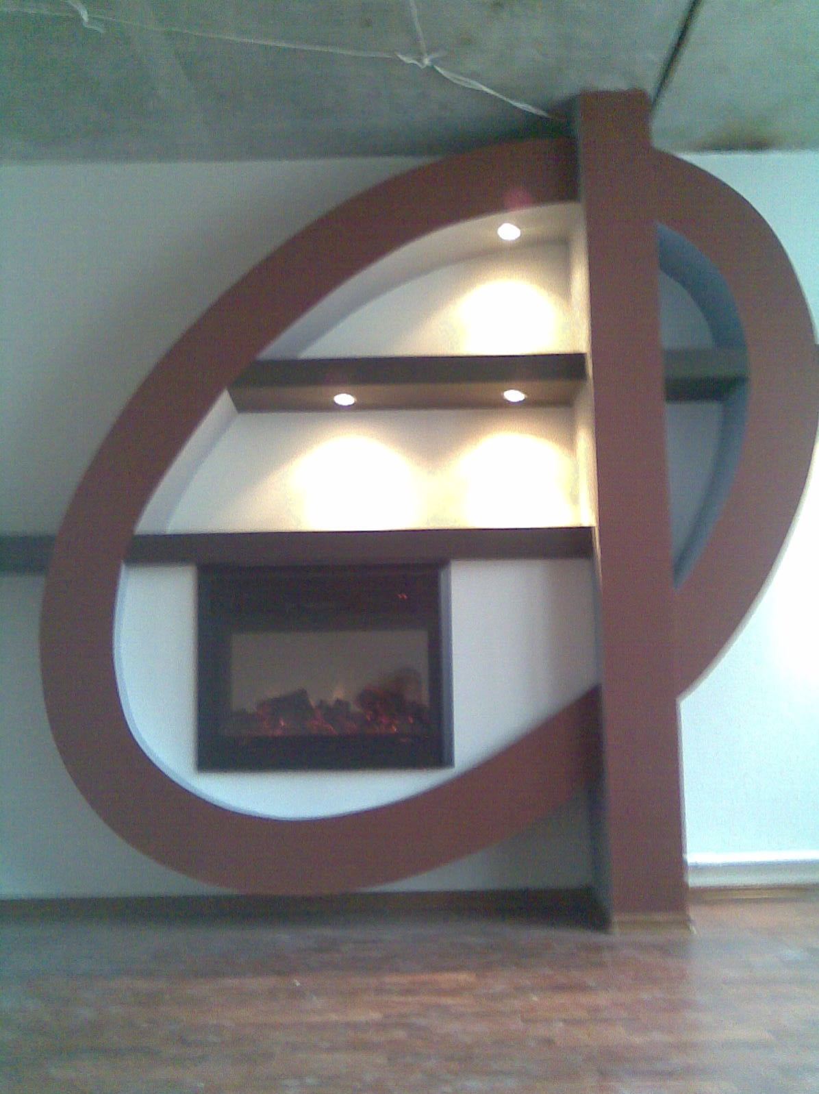 Nietypowa zabudowa kominka elektrycznego z półkami i oświetleniem punktowym, brązowa rama w kształcie elipsy, surowy betonowy sufit w tle.