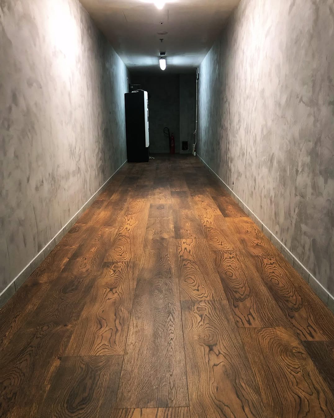 Długi korytarz z nową, ciemną, drewnianą podłogą i szarymi ścianami o fakturze betonu, zakończony automatem z napojami i gaśnicą w tle.