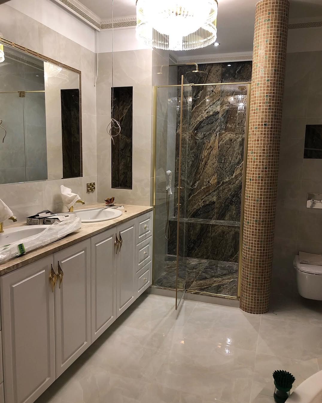 Luksusowa łazienka w trakcie wykańczania: białe szafki z ozdobnymi uchwytami, marmurowy blat, kabina prysznicowa z kamienną ścianą, kolumna z mozaiki i kryształowy żyrandol