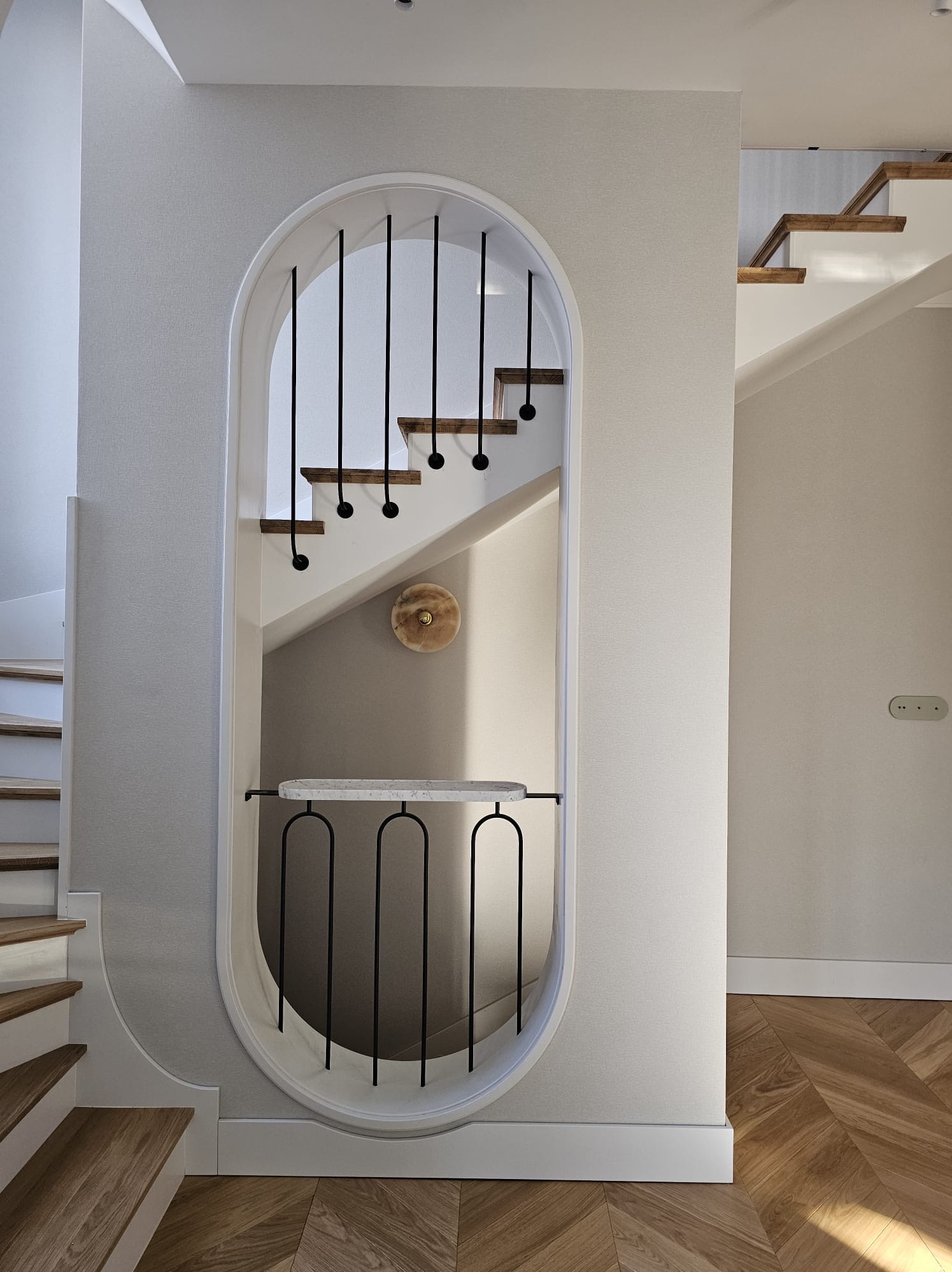 Nowoczesne schody z drewnianymi stopniami i czarnymi balustradami, wkomponowane w owalną niszę z marmurowym blatem i oświetleniem ściennym, parkiet w jodełkę na podłodze.