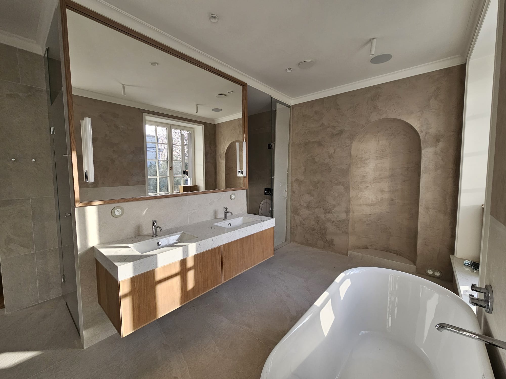 Nowoczesna łazienka z podwójną umywalką na drewnianej szafce z blatem z białego kamienia, duże lustro w drewnianej ramie, kabina prysznicowa ze szklanymi drzwiami, wolnostojąca biała wanna i ściany...