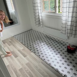 Krystian Rapacz - Przygotowanie podłoża pod panele podłogowe z widoczną folią izolacyjną, odbicie uśmiechniętej dziewczynki w lustrze, okno z białą ramą i zasłonami w kratę.