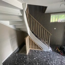 Krystian Rapacz - Białe, kręcone schody z drewnianą balustradą w trakcie montażu, widoczne narzędzia na podłodze o wzorze lastryko, ściana wykończona pionowymi panelami drewnianymi.