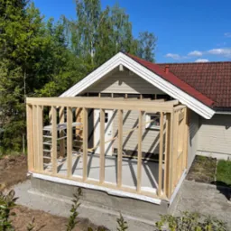 Drewniana konstrukcja dobudowy do domu jednorodzinnego z widoczną drabinką, fundamentem i częścią elewacji w słoneczny dzień.