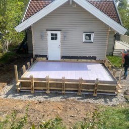 Krystian Rapacz - Przygotowanie fundamentów pod taras przy szarym domu z czerwoną dachówką, widoczne drewniane szalunki i izolacja z różowego styropianu, obok leżące deski i pracownik w tle.