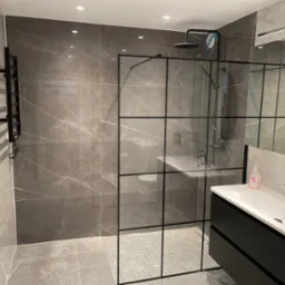 Nowoczesna łazienka z kabiną prysznicową typu walk-in z czarnymi szprosami, szare płytki imitujące marmur na ścianach i podłodze, czarna szafka pod umywalką z białą umywalką nablatową, grzejnik...