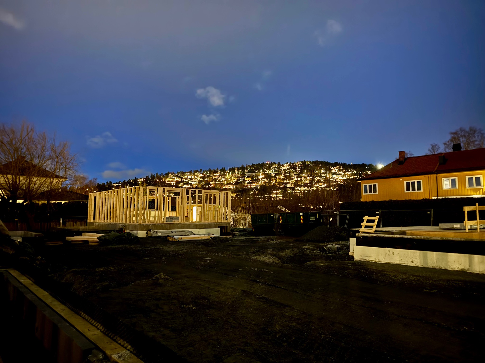 Nocny widok na plac budowy z drewnianą konstrukcją domu w trakcie wznoszenia, na tle oświetlonego wzgórza zabudowanego domami.