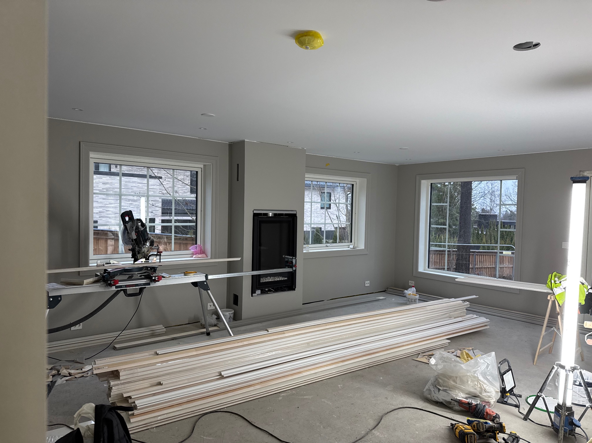 Wnętrze w trakcie prac wykończeniowych: ułożone listwy przypodłogowe, piła ukosowa na stole, narzędzia budowlane na podłodze, widok przez okno na domy.