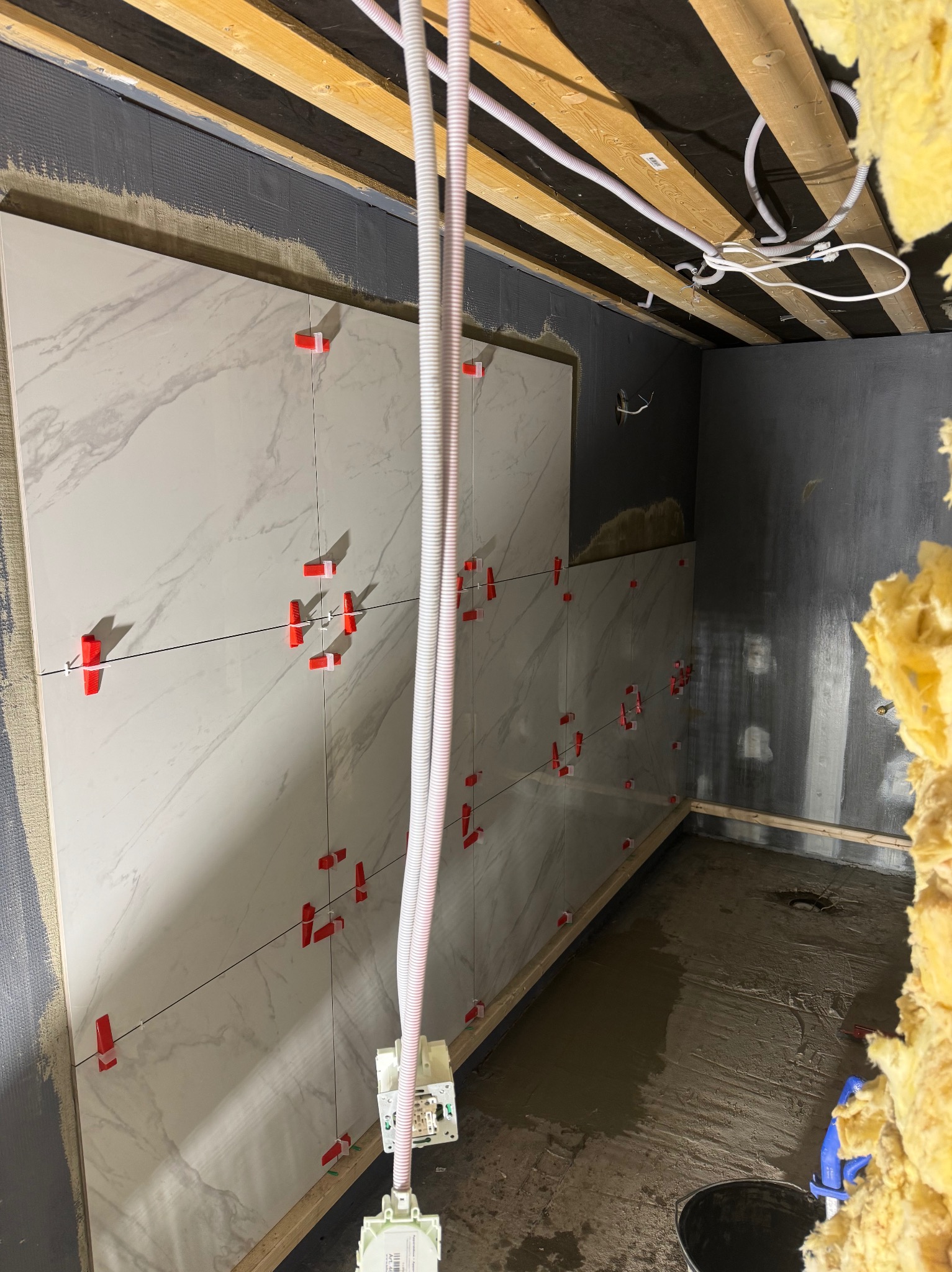 Montaż płytek imitujących marmur na ścianie z użyciem czerwonych krzyżyków dystansowych, widoczne przewody elektryczne i surowy stan wykończenia pomieszczenia.