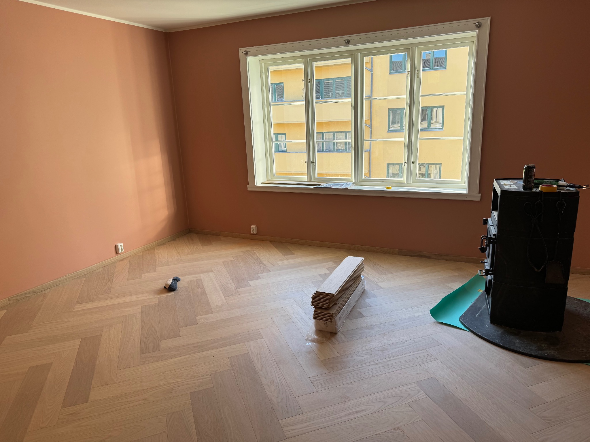 Jasny parkiet ułożony we wzór jodełki francuskiej w pokoju z łososiowymi ścianami, z oknem wychodzącym na żółty budynek, paczka paneli i piec na drewno w rogu.