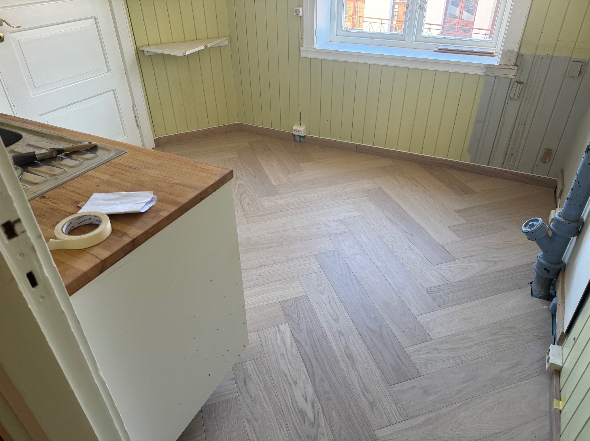 Układanie jasnego parkietu w jodełkę francuską w kuchni z żółtymi panelami ściennymi, widoczne narzędzia i taśma malarska na blacie.