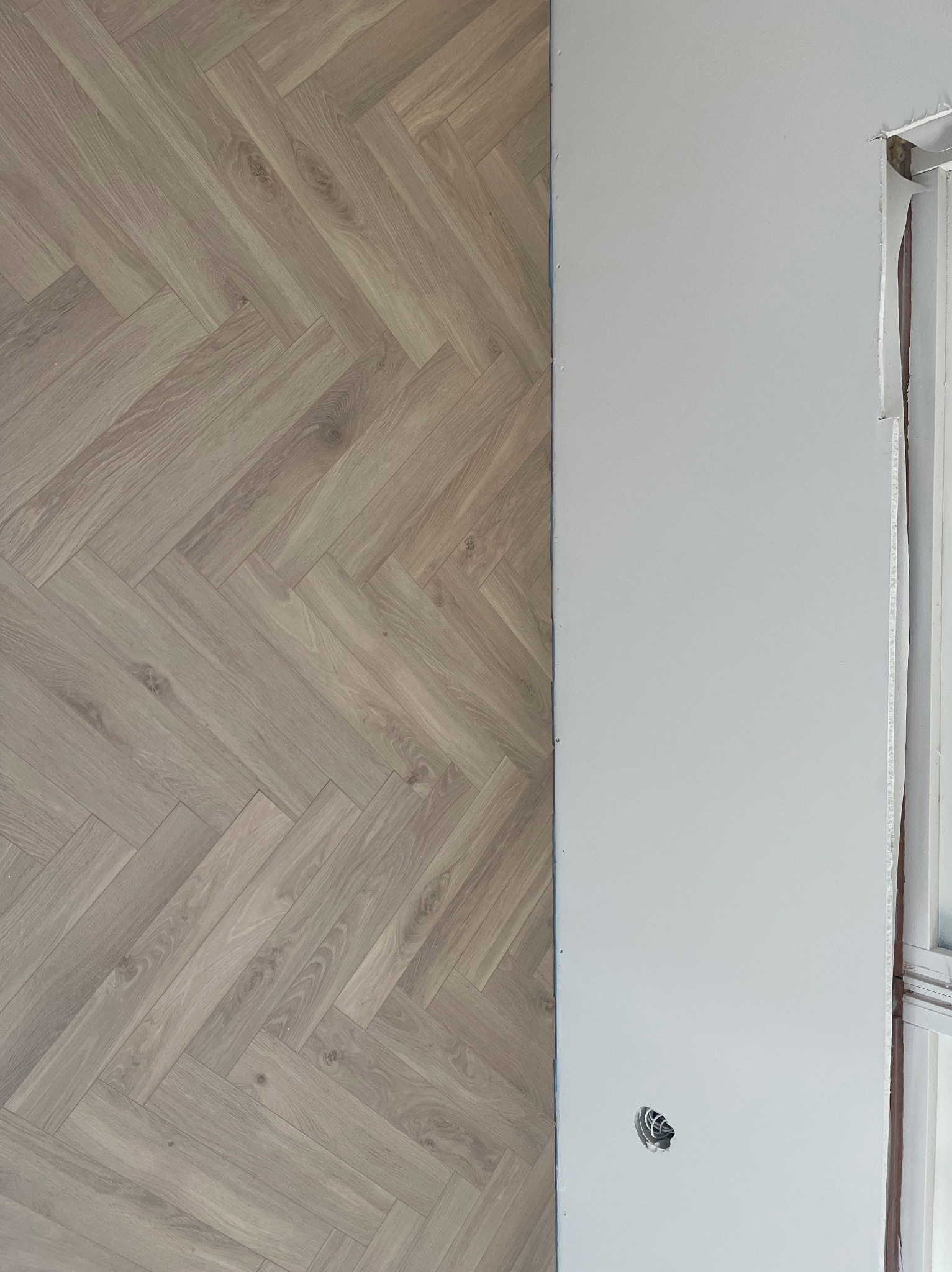 Fragment ściany z ułożonym parkietem w jodełkę i częściowo zamontowaną płytą karton gips, widoczne gniazdko elektryczne w ścianie.