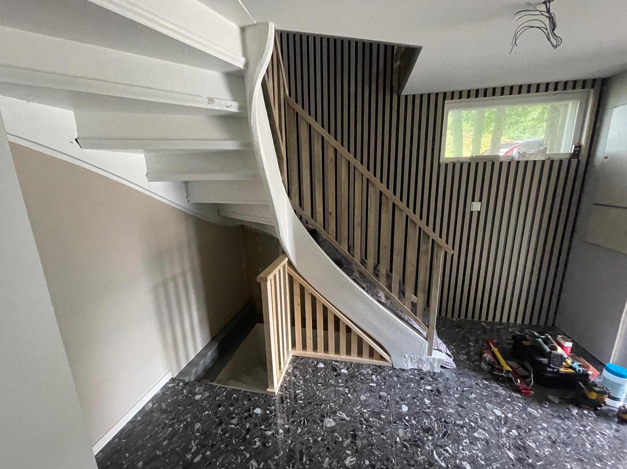 Białe, kręcone schody z drewnianą balustradą w trakcie montażu, widoczne narzędzia na podłodze o wzorze lastryko, ściana wykończona pionowymi panelami drewnianymi.