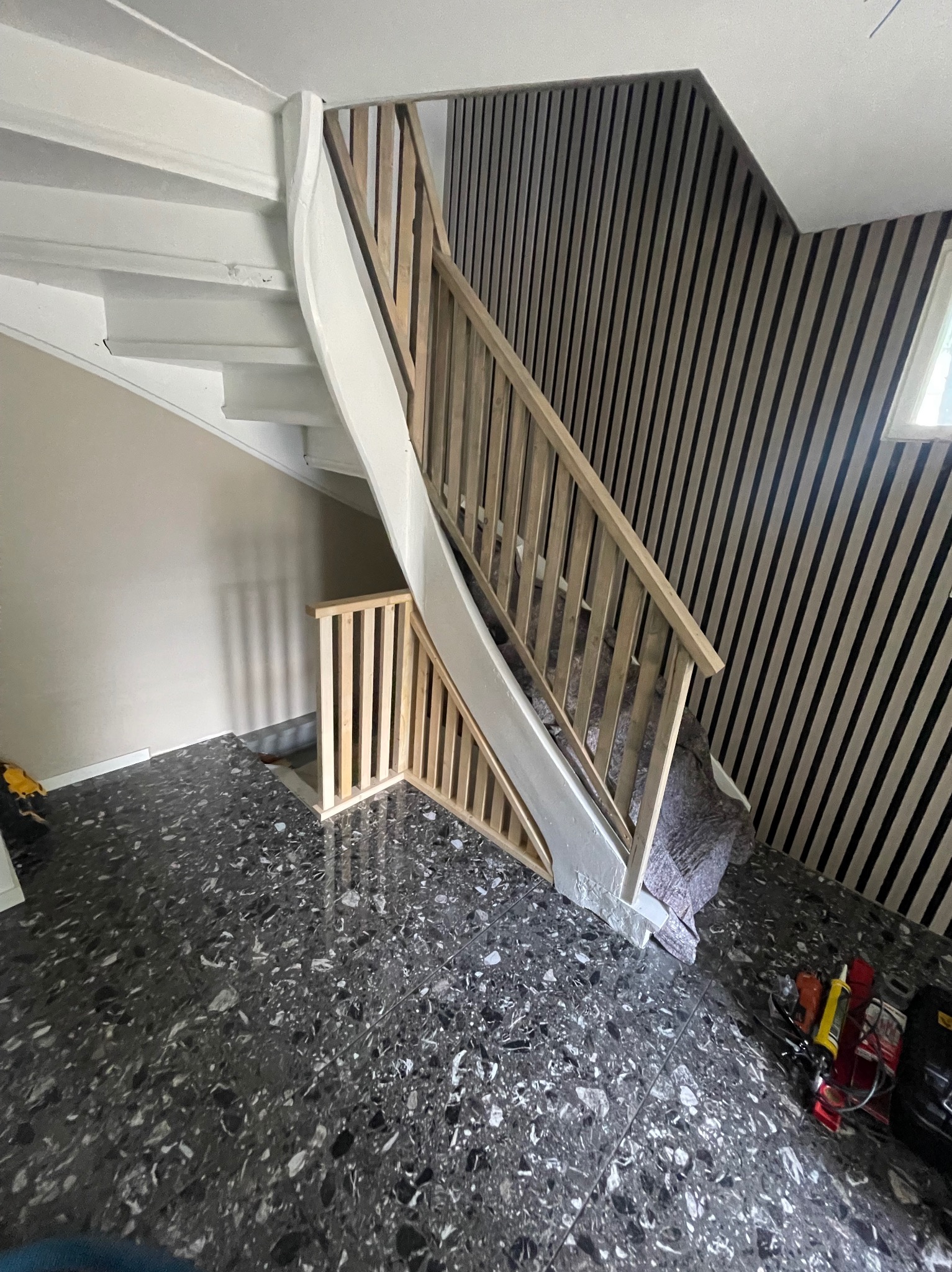 Montaż drewnianej balustrady schodowej z białymi elementami, widoczne narzędzia i zabezpieczenia na podłodze z lastryko, ściana wykończona panelami pionowymi.