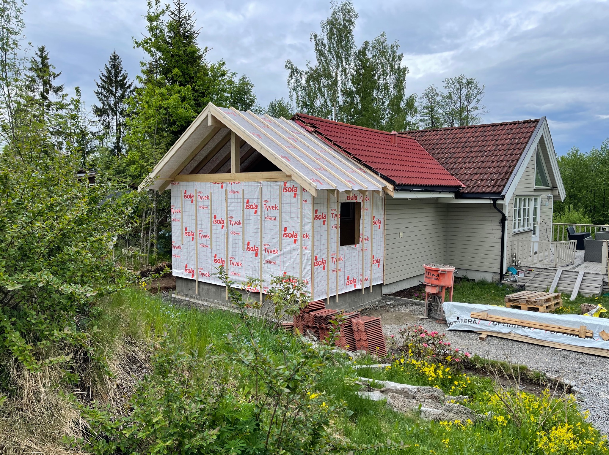 Budowa dobudówki do domu jednorodzinnego w trakcie realizacji, widoczne drewniane elementy konstrukcji dachu pokryte membraną izolacyjną, w tle istniejąca część domu z czerwoną dachówką...