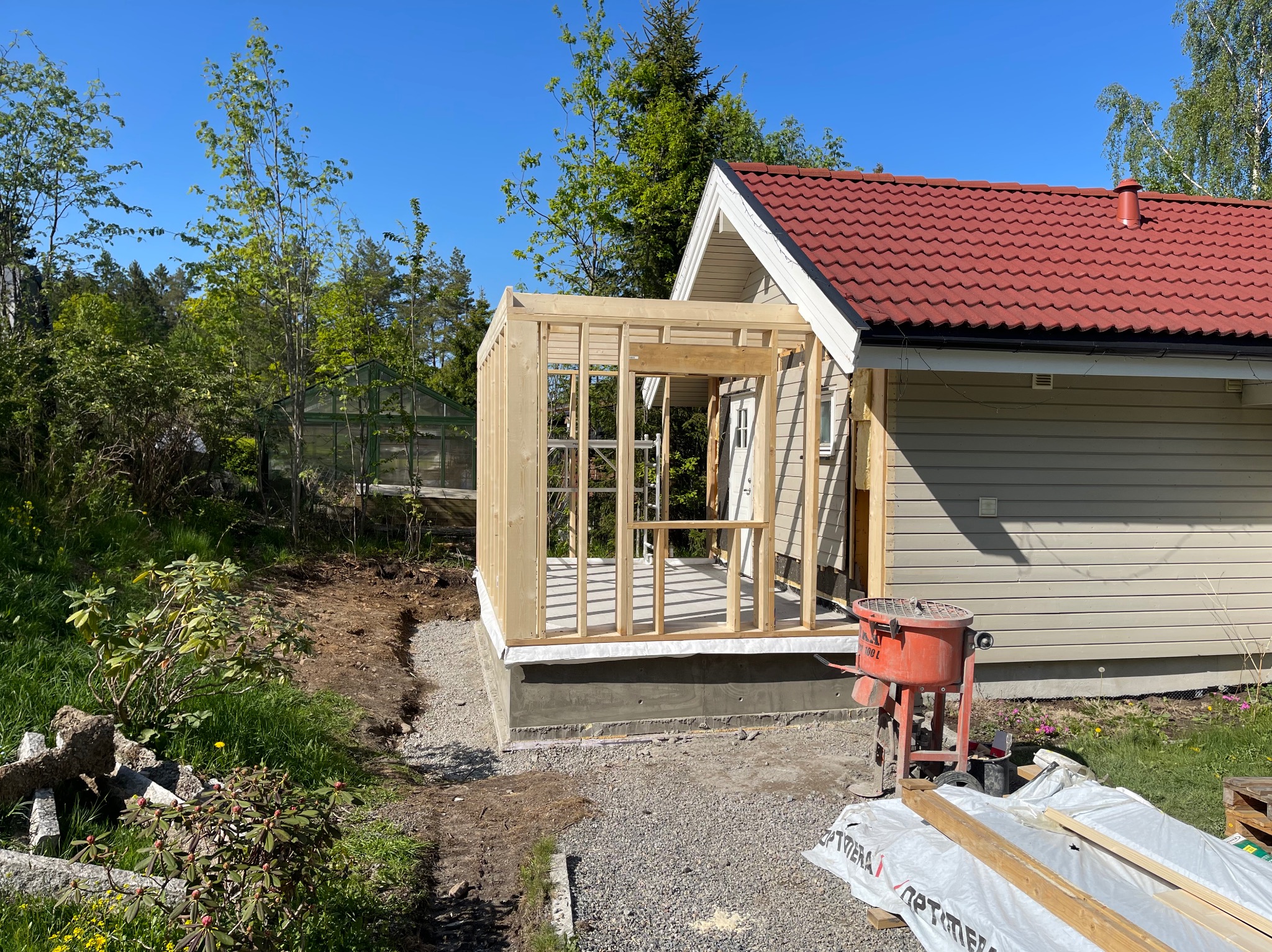 Drewniana konstrukcja dobudowy do domu z czerwonym dachem, widoczny mikser do betonu, teren w trakcie przygotowania, w tle szklarnia.