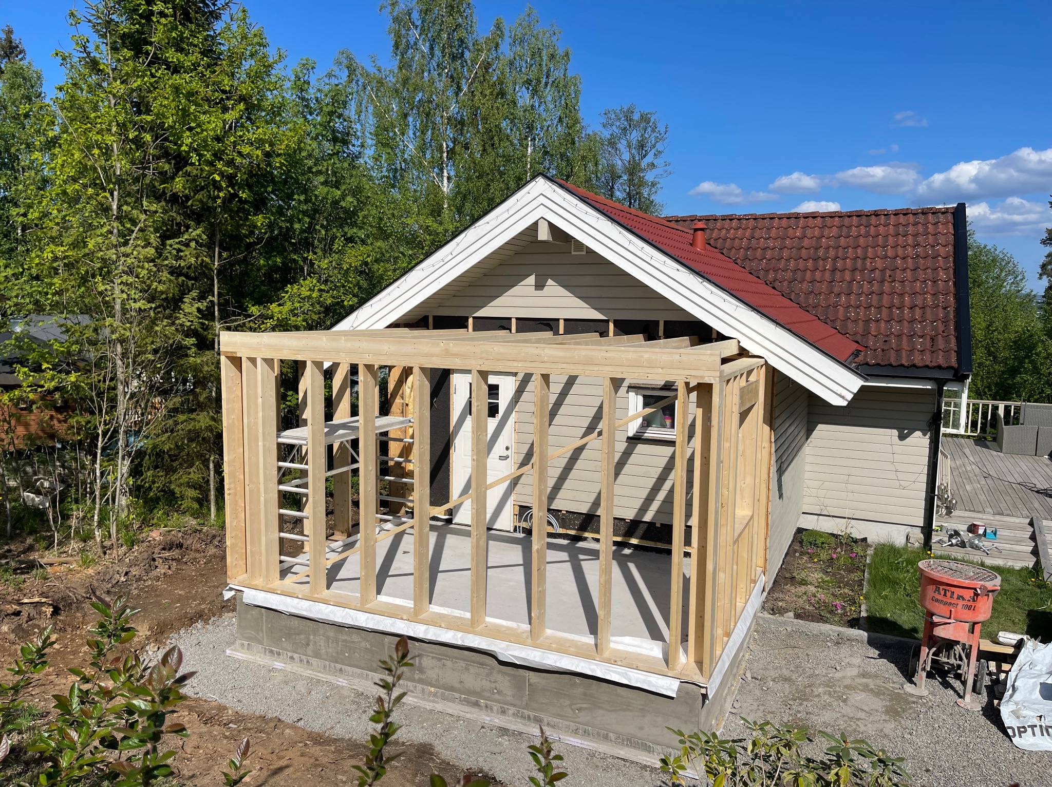 Drewniana konstrukcja dobudowy do domu jednorodzinnego z widoczną drabinką, fundamentem i częścią elewacji w słoneczny dzień.