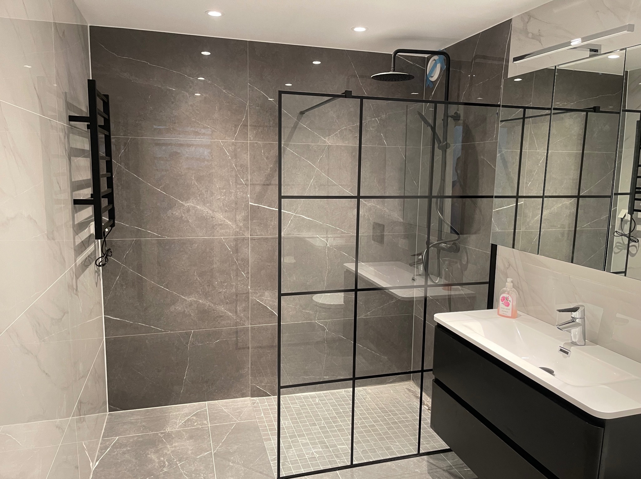 Nowoczesna łazienka z kabiną prysznicową typu walk-in z czarnymi szprosami, szare płytki imitujące marmur na ścianach i podłodze, czarna szafka pod umywalką z białą umywalką nablatową, grzejnik...
