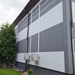 Mariusz Kamiński - Elewacja budynku z poziomymi pasami szarości i bieli, z widocznymi rurami spustowymi i dwoma klimatyzatorami na ścianie. Obok rośnie zielone drzewo.