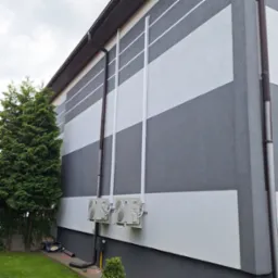 Elewacja budynku z poziomymi pasami szarości i bieli, z widocznymi rurami spustowymi i dwoma klimatyzatorami na ścianie. Obok rośnie zielone drzewo.