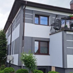 Mariusz Kamiński - Dwukondygnacyjny dom z elewacją w odcieniach szarości i bieli, z balkonem z kutą balustradą i brązowymi oknami, otoczony zielenią.