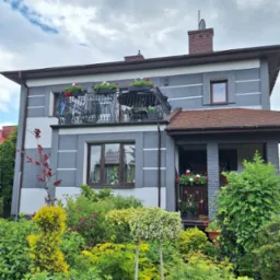 Dwukondygnacyjny dom z szarą elewacją i dekoracyjnymi listwami. Balkon z kutą balustradą i kwiatami. Bujna roślinność przed budynkiem. Dachówka w kolorze czerwonej cegły.