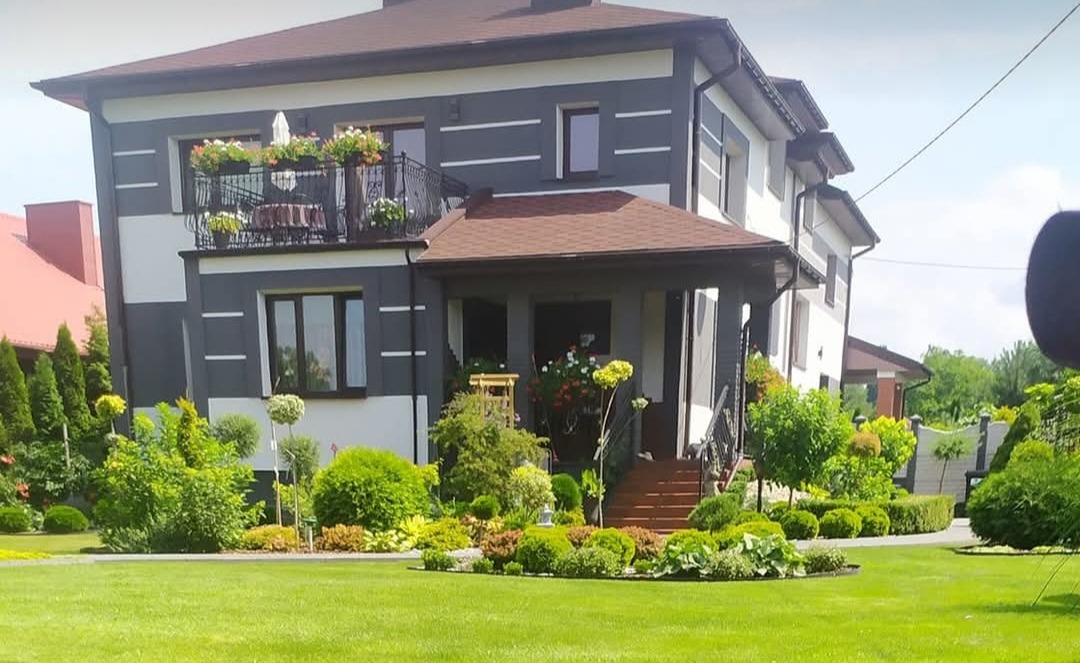 Dwukondygnacyjny dom z szarą elewacją i białymi akcentami, balkon z kwiatami, zadbany ogród z trawnikiem i ozdobnymi krzewami, wejście z drewnianymi schodami.