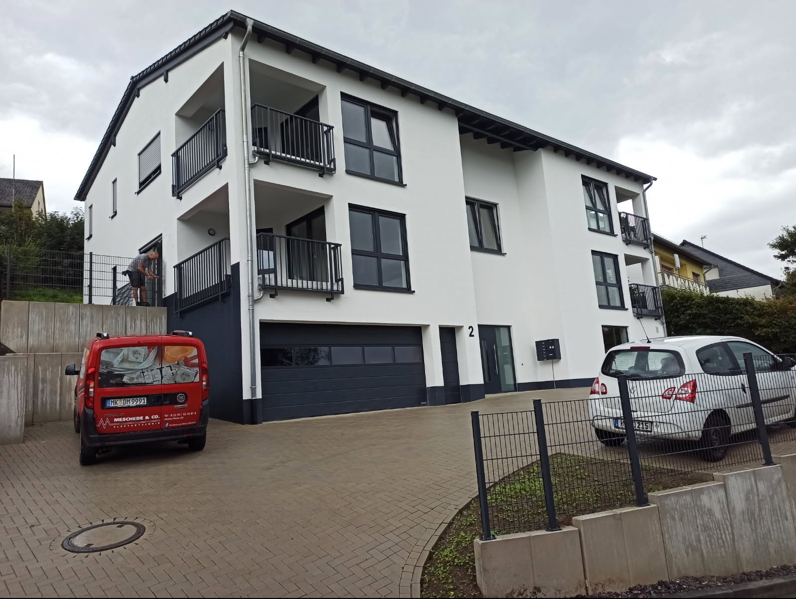 Nowoczesny, trzypiętrowy budynek mieszkalny w Sochaczewie z białą elewacją, czarnymi oknami i balkonami. Na balkonie widoczna osoba w trakcie pracy. Na podjeździe zaparkowane dwa samochody.