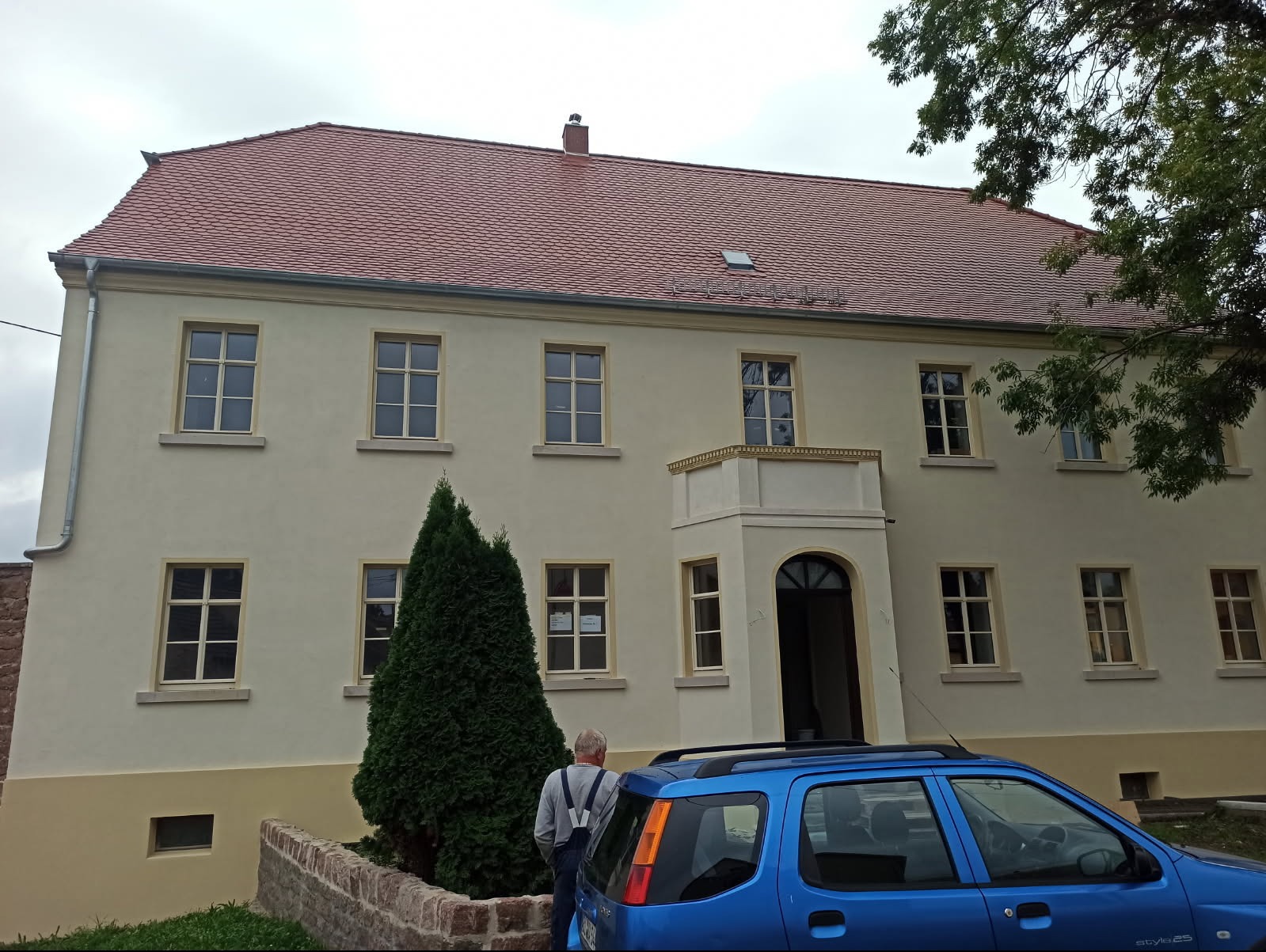 Odnowiona elewacja budynku z beżowym tynkiem, czerwonym dachem i jasnymi obramowaniami okien, widoczny fragment niebieskiego samochodu i sylwetka mężczyzny z szelkami.