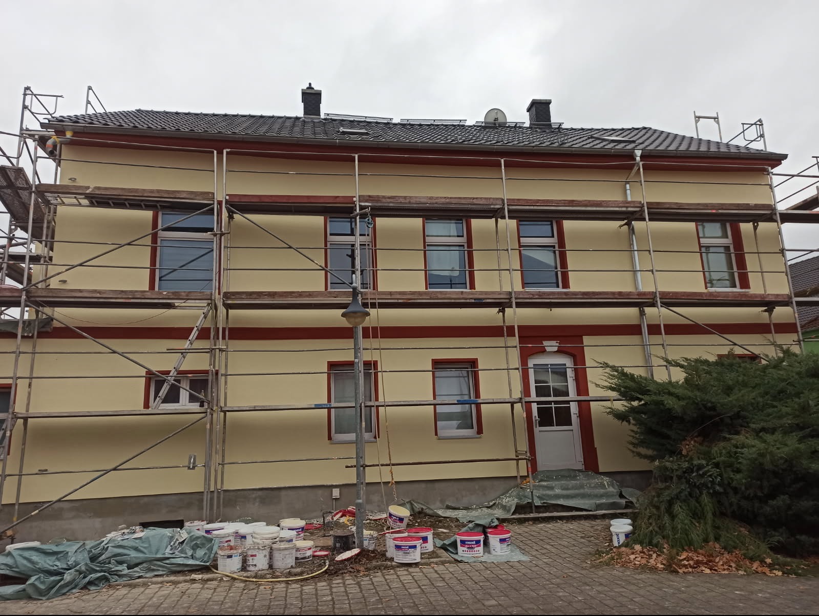 Dwupiętrowy budynek z elewacją w odcieniach beżu i czerwieni, otoczony rusztowaniami; widoczne puszki farb i narzędzia malarskie u podstawy budynku, sugerujące prace wykończeniowe.
