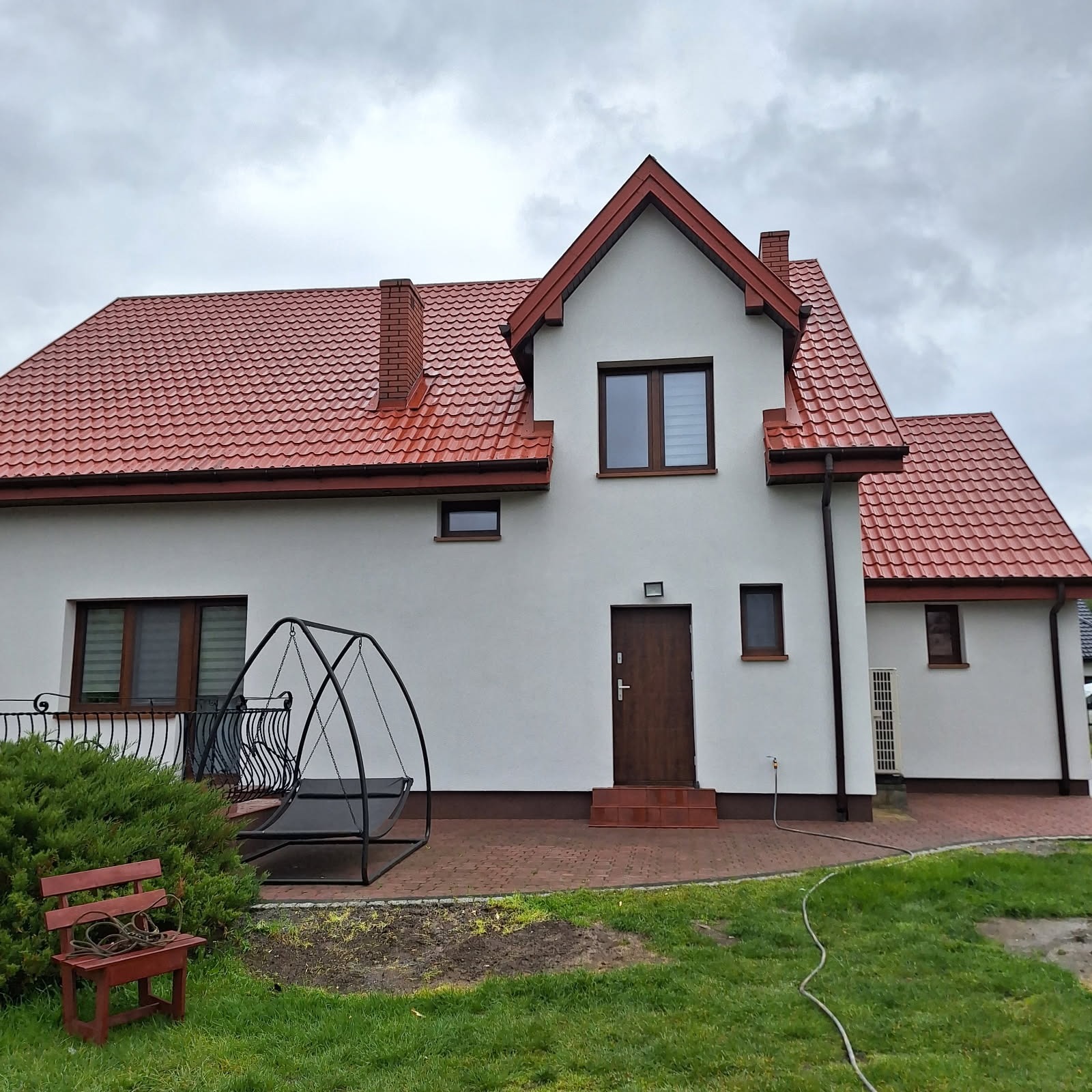 Dom jednorodzinny z białą elewacją i czerwoną dachówką, widok z ogrodu. Na pierwszym planie huśtawka i ławka, w tle fragment elewacji z oknami i drzwiami wejściowymi.