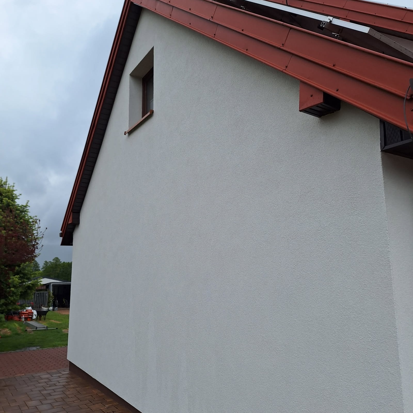 Boczny widok otynkowanej, jasnoszarej elewacji domu z brązowym dachem i oknem, na tle pochmurnego nieba i zieleni ogrodu. Widoczny fragment chodnika z kostki.