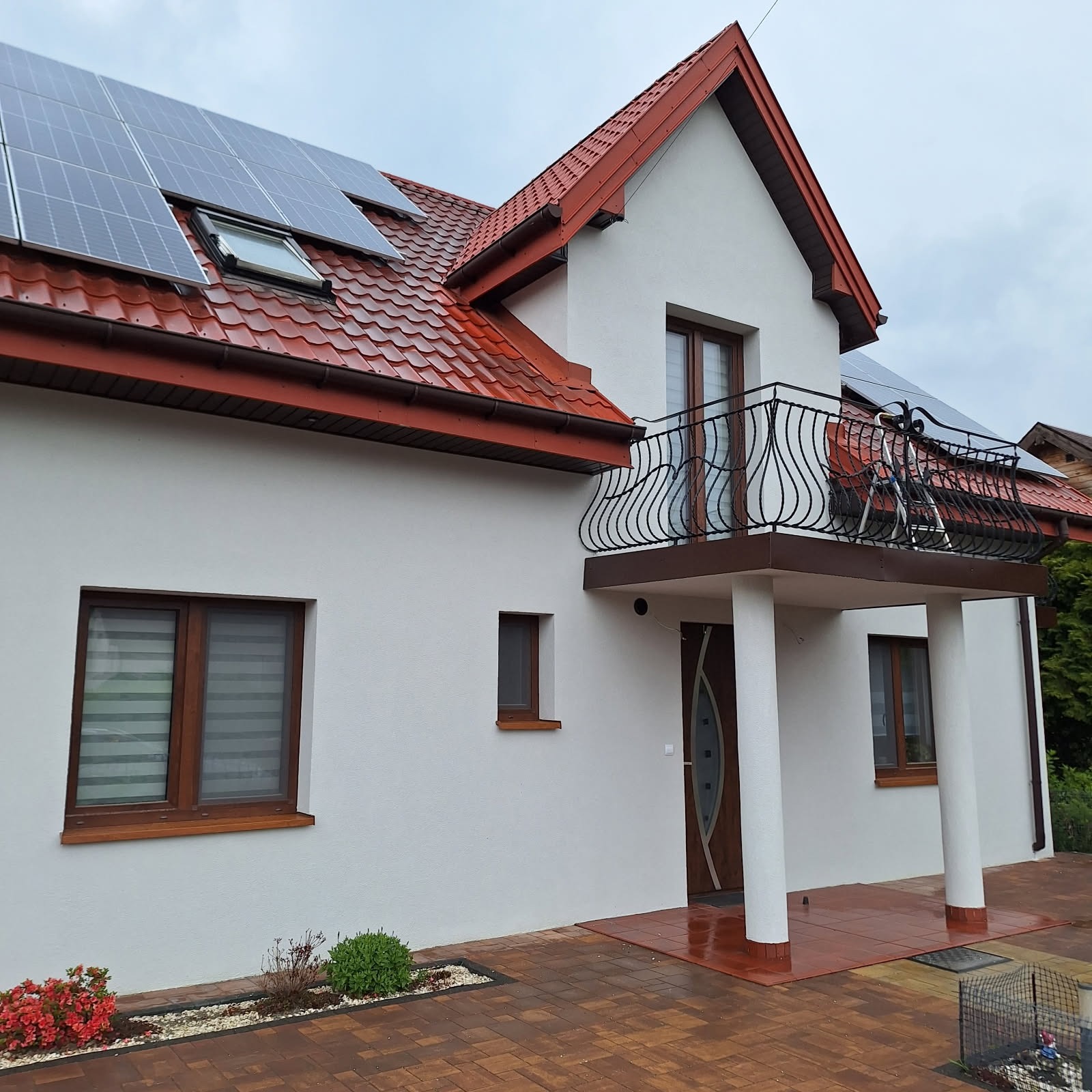 Biały dom z czerwoną dachówką i panelami słonecznymi. Balustrada balkonu z ozdobnymi, czarnymi elementami. Widoczne okna z drewnianą stolarką i brązowa kostka brukowa.
