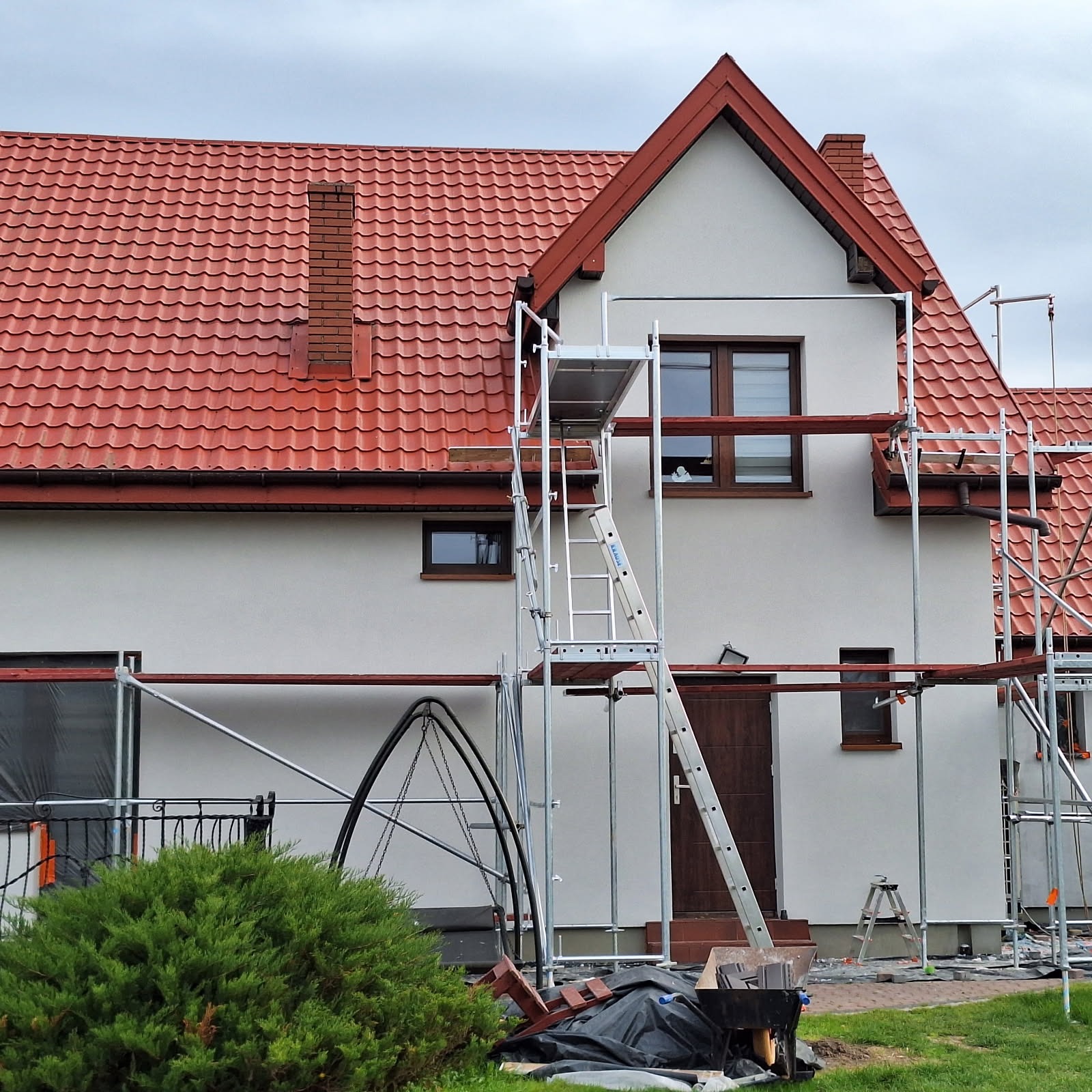 Dom z czerwoną dachówką w trakcie prac elewacyjnych. Widoczne rusztowania, okna z brązowymi ramami i fragment zielonego krzewu w dolnej części kadru.