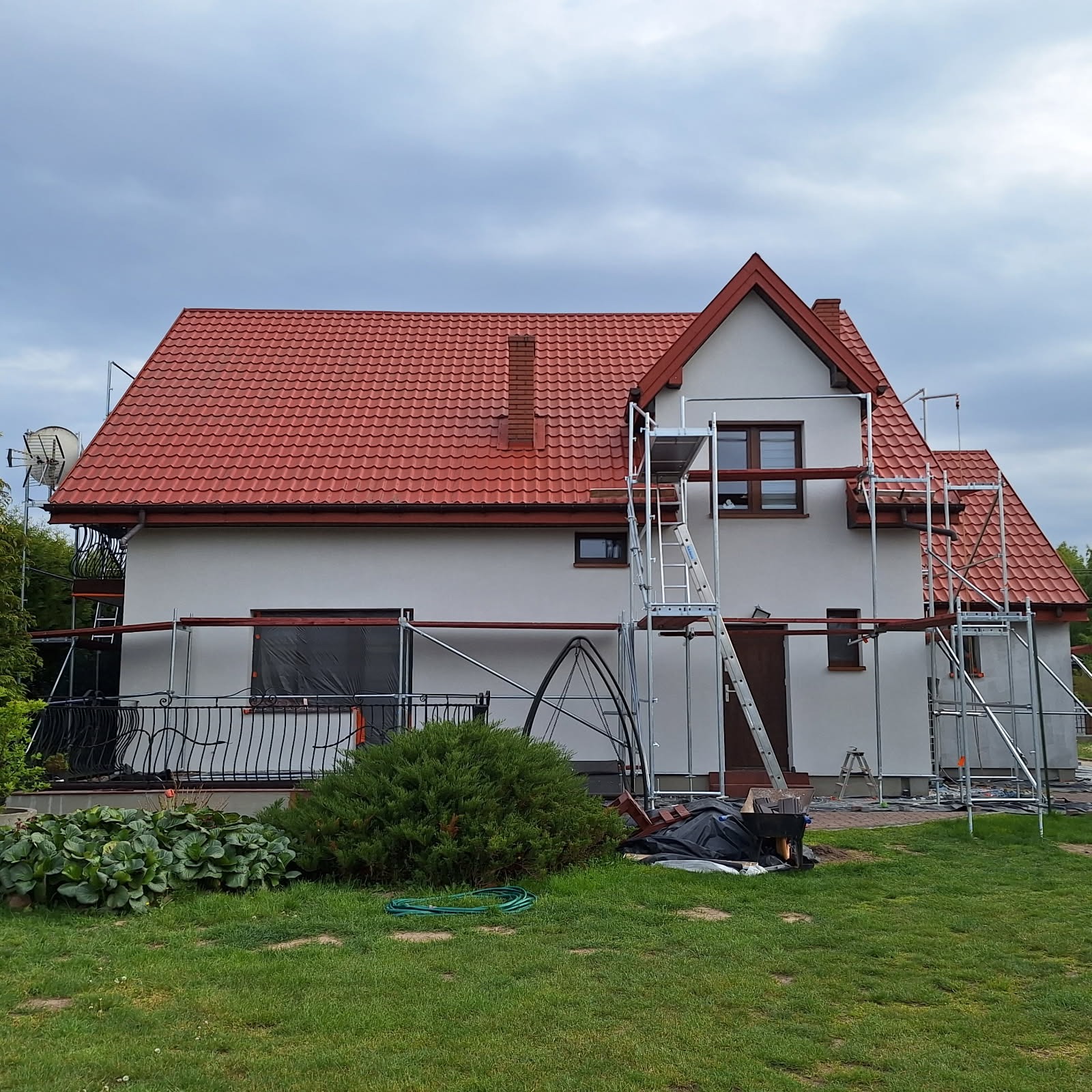 Dom jednorodzinny w trakcie prac elewacyjnych, rusztowania, zabezpieczone okno folią, czerwony dach i biała elewacja. Widok z poziomu trawnika.