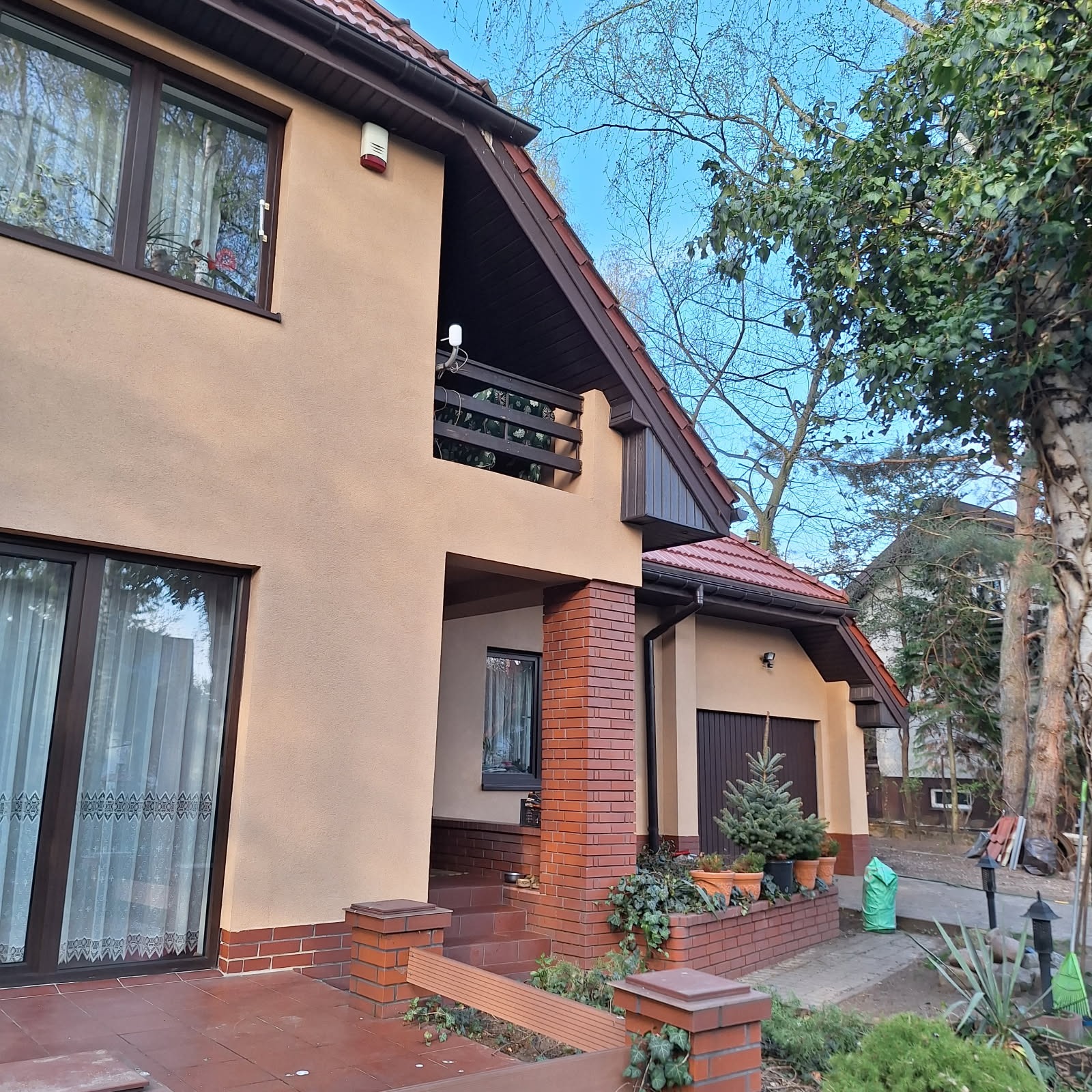 Dom jednorodzinny z beżową elewacją i balkonem, z elementami wykończenia z cegły. Widoczne drzewa, fragment chodnika i roślinność w otoczeniu budynku.