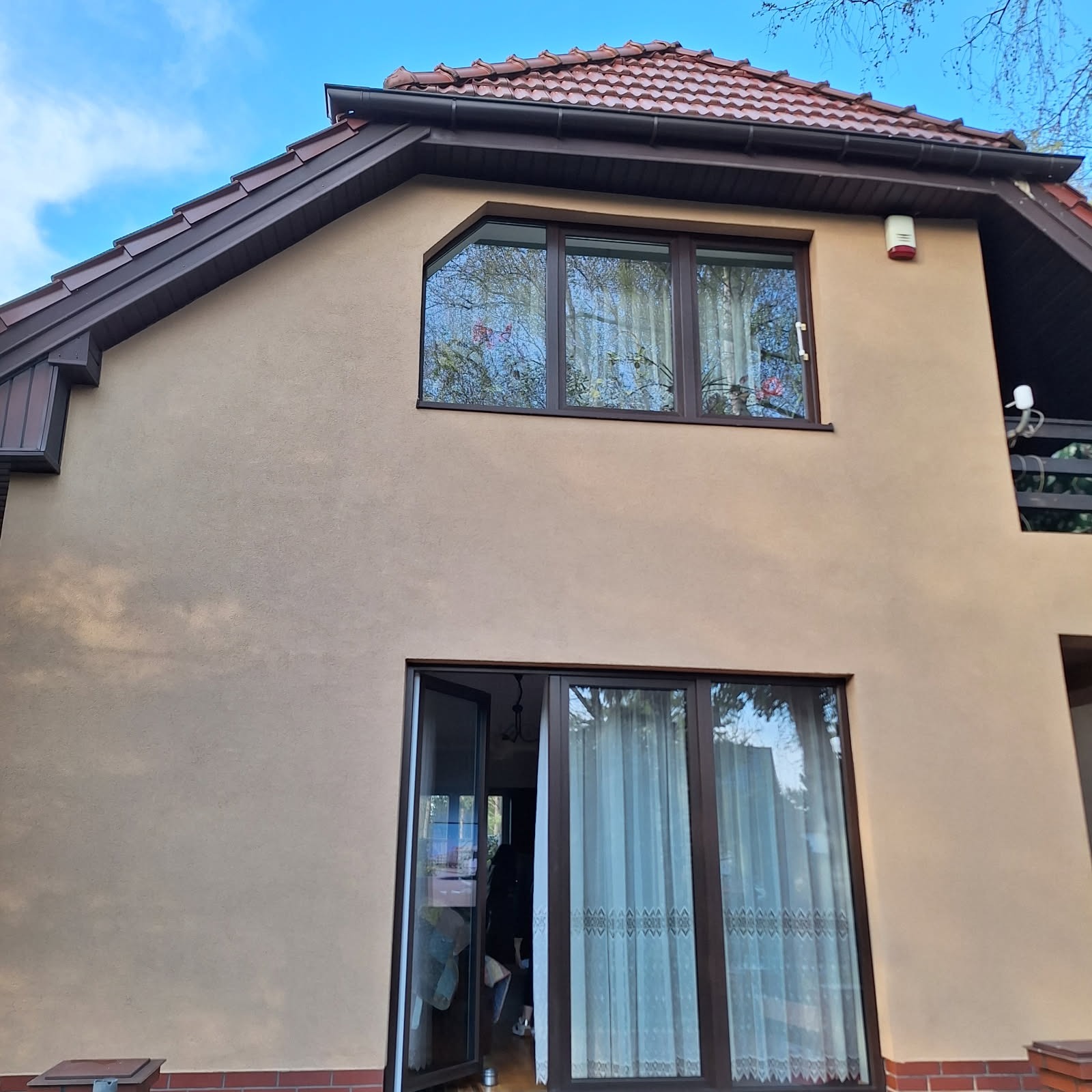 Dwupiętrowy dom z beżową elewacją, brązową stolarką okienną i czerwoną dachówką. Widoczne okna z firankami i balkon.