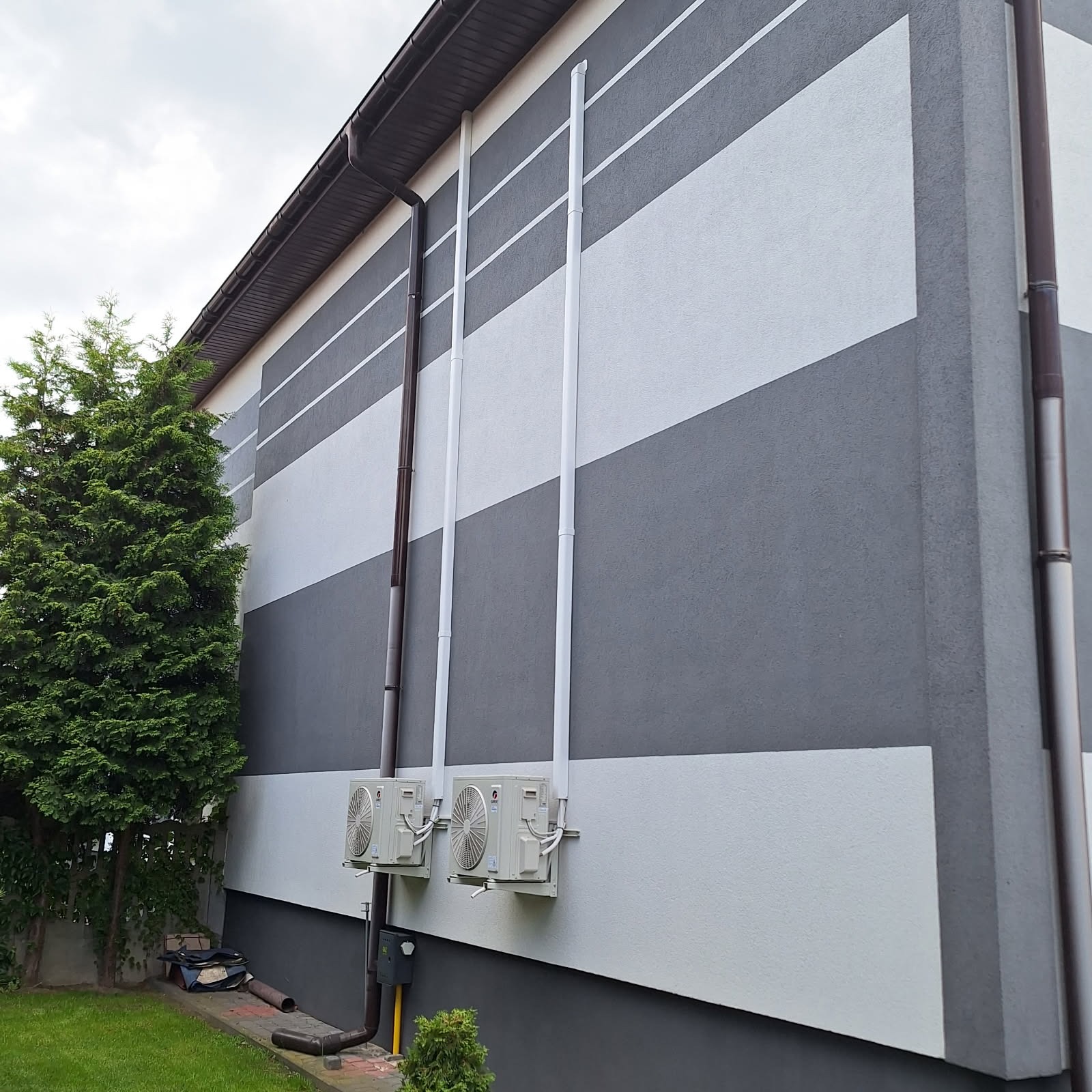 Elewacja budynku z poziomymi pasami szarości i bieli, z widocznymi rurami spustowymi i dwoma klimatyzatorami na ścianie. Obok rośnie zielone drzewo.