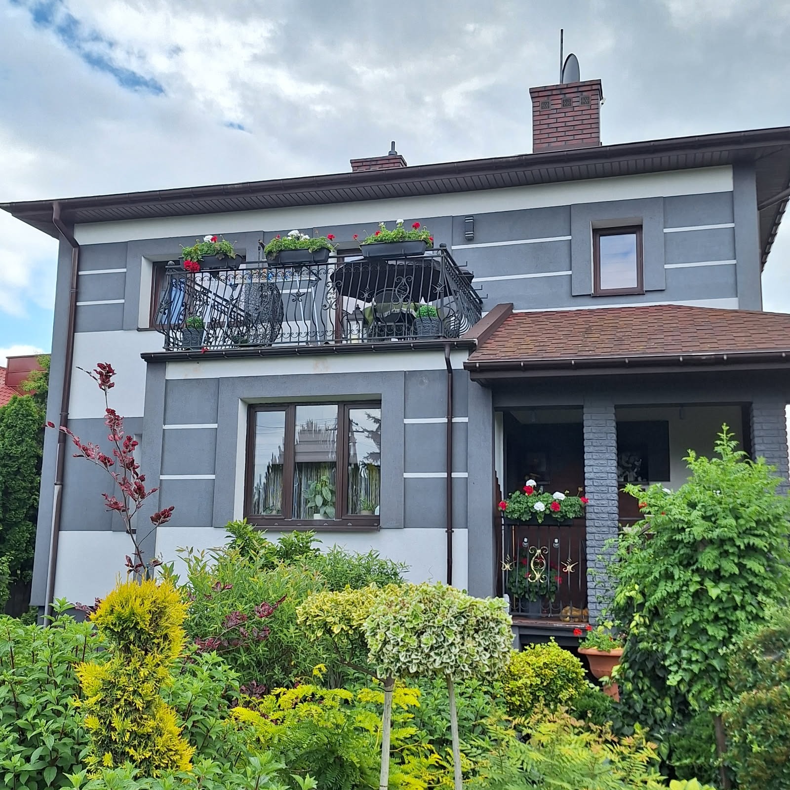 Dwukondygnacyjny dom z szarą elewacją i dekoracyjnymi listwami. Balkon z kutą balustradą i kwiatami. Bujna roślinność przed budynkiem. Dachówka w kolorze czerwonej cegły.