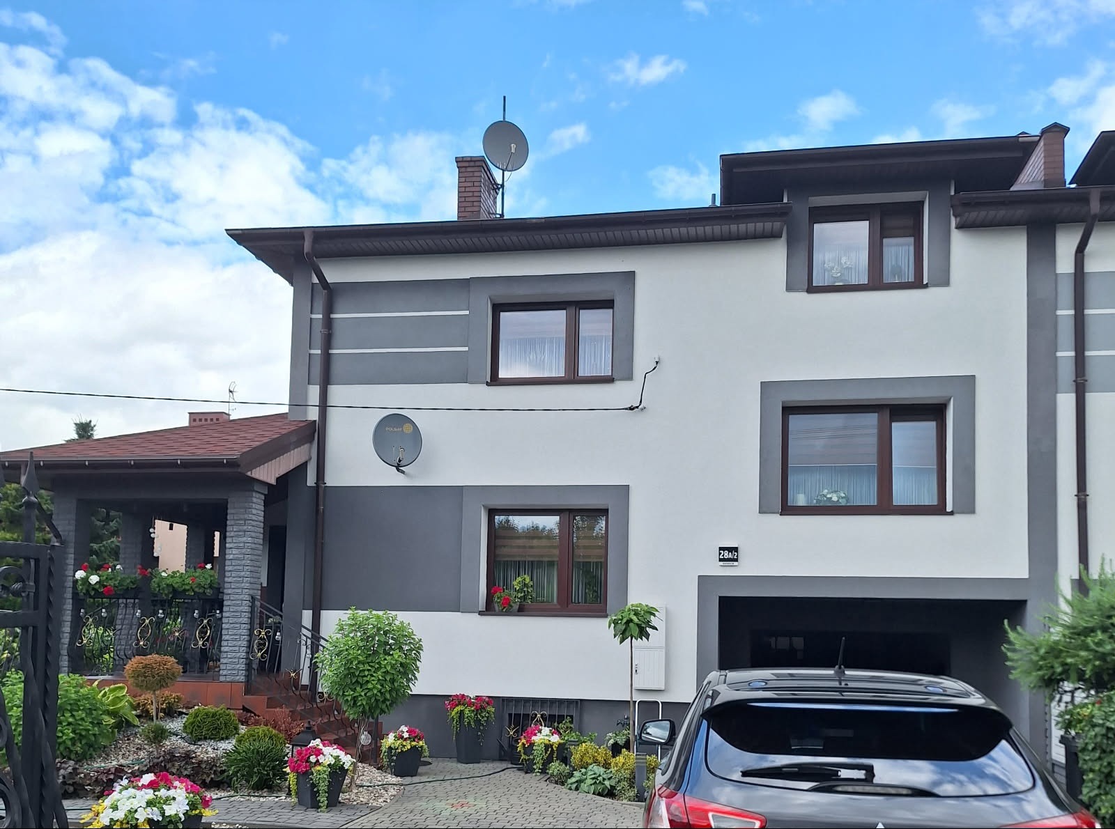 Dwukondygnacyjny dom z szarobiałą elewacją, brązową stolarką okienną i satelitami. Ogród z kwiatami i krzewami. Błękitne niebo z chmurami w tle.