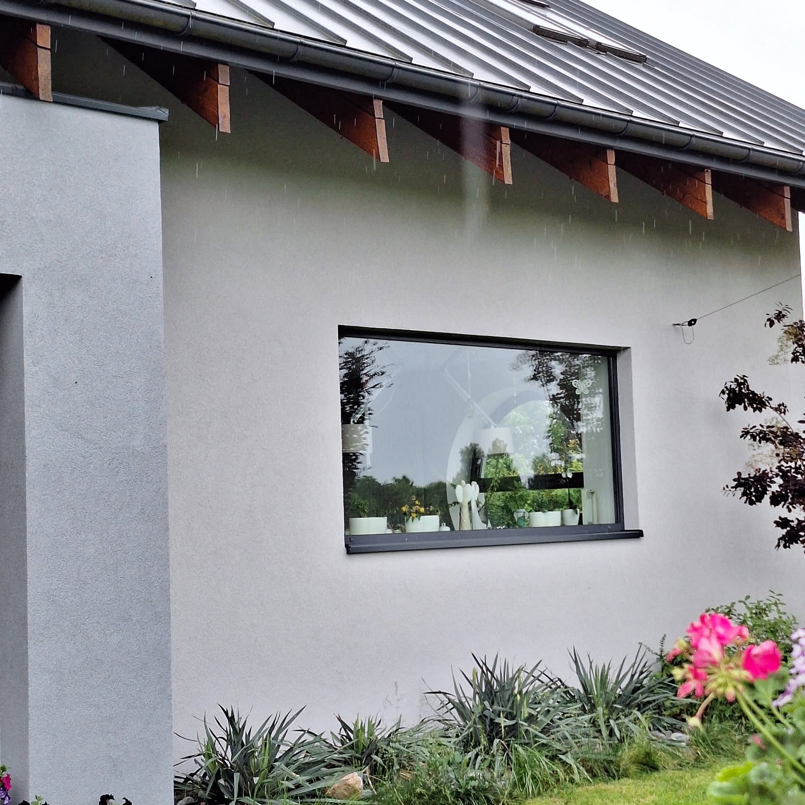 Jasnoszara elewacja domu jednorodzinnego z dużym oknem, w tle widoczne krople deszczu i fragment metalowego dachu. Wokół roślinność.