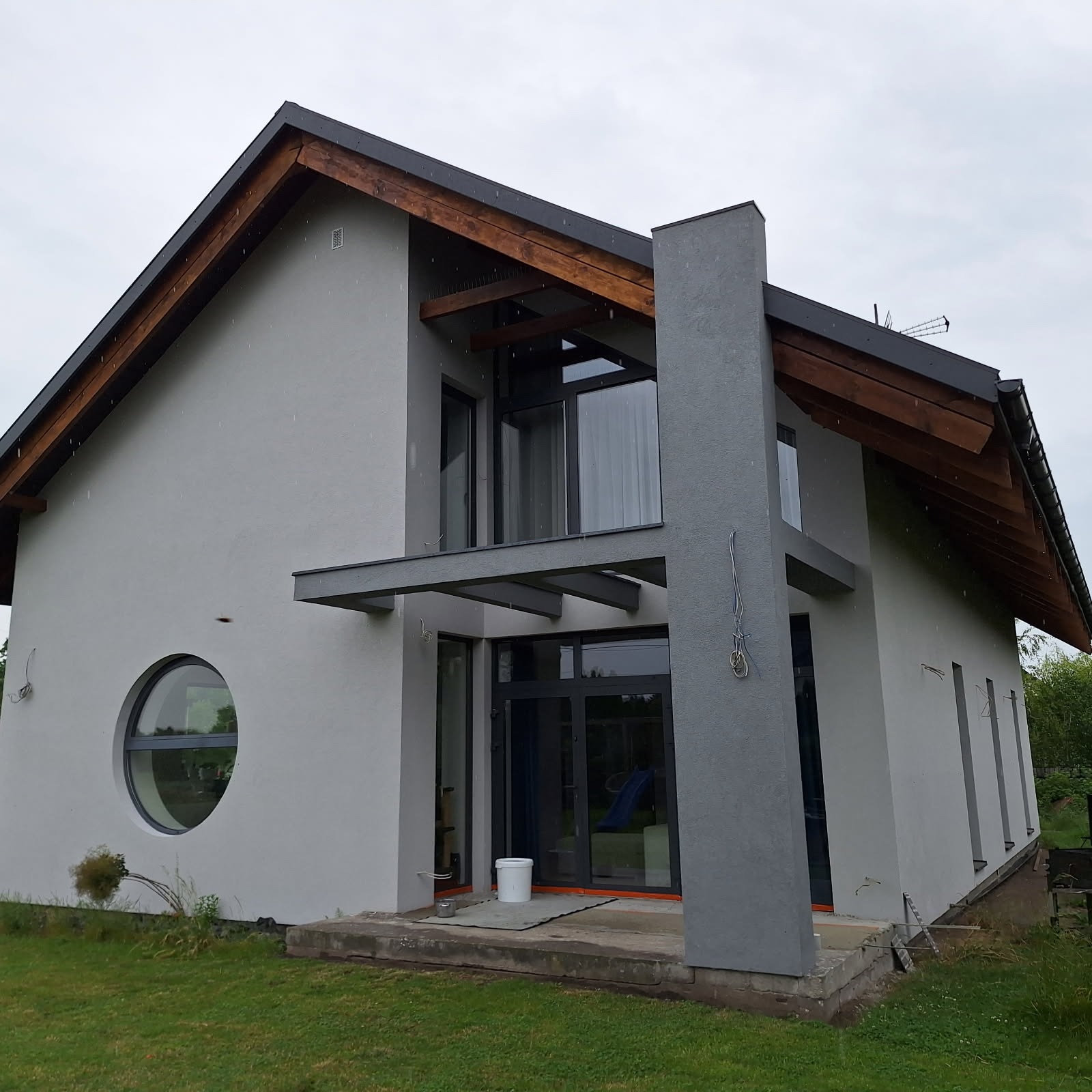 Nowoczesny dom z jasnoszarą elewacją, okrągłym oknem i drewnianym zadaszeniem. Widoczne ślady po pracach elewacyjnych i narzędzia na tarasie. Pochmurny dzień.