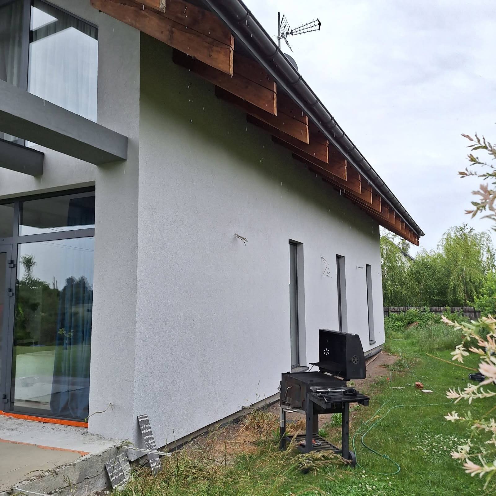 Boczny widok otynkowanej, jasnoszarej elewacji domu z drewnianymi elementami okapu, anteną i grillem w ogrodzie, w pochmurny dzień.