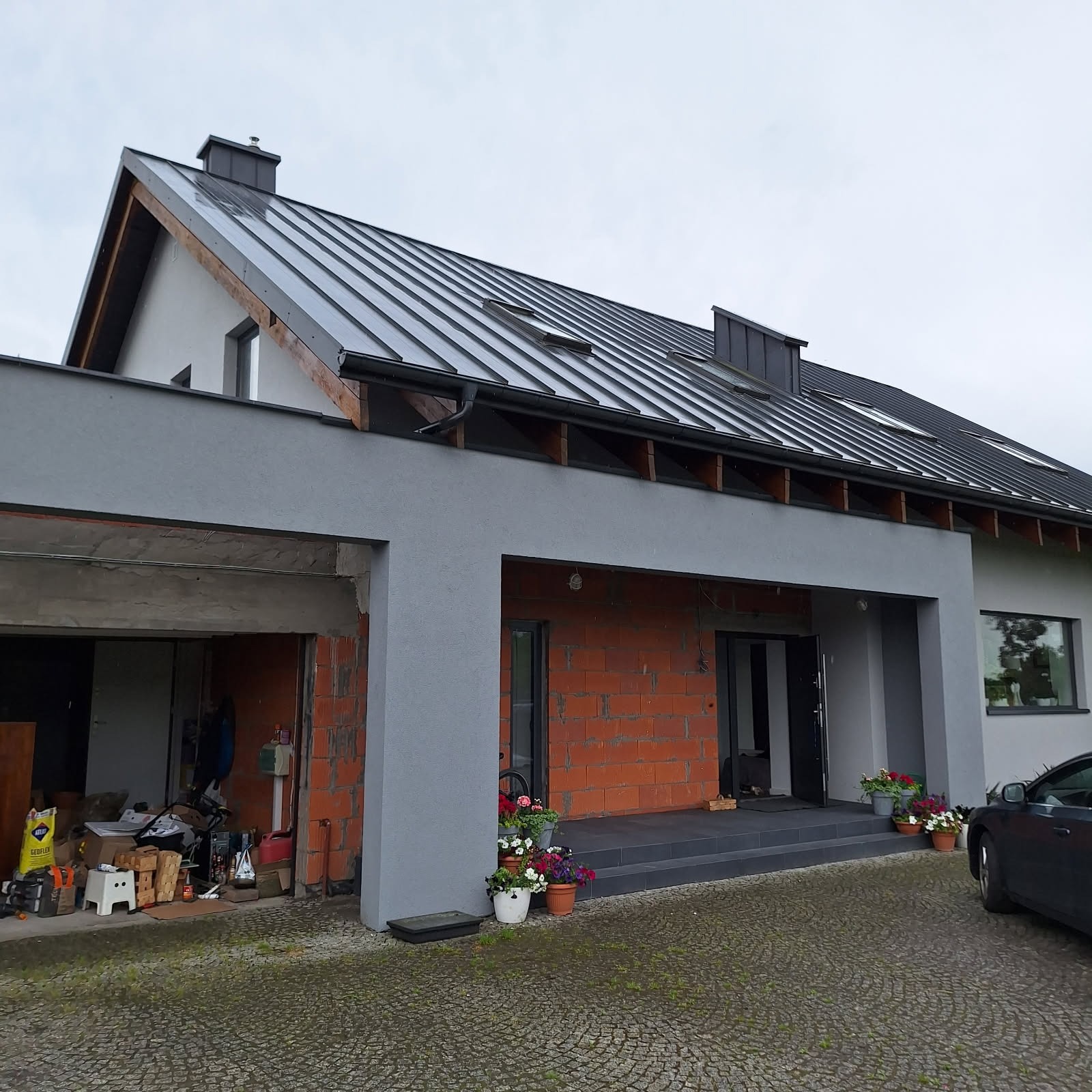 Dom z szarą elewacją i metalowym dachem w trakcie budowy, widoczne cegły i kwiaty w donicach. Częściowo wykończony budynek z garażem i samochodem.
