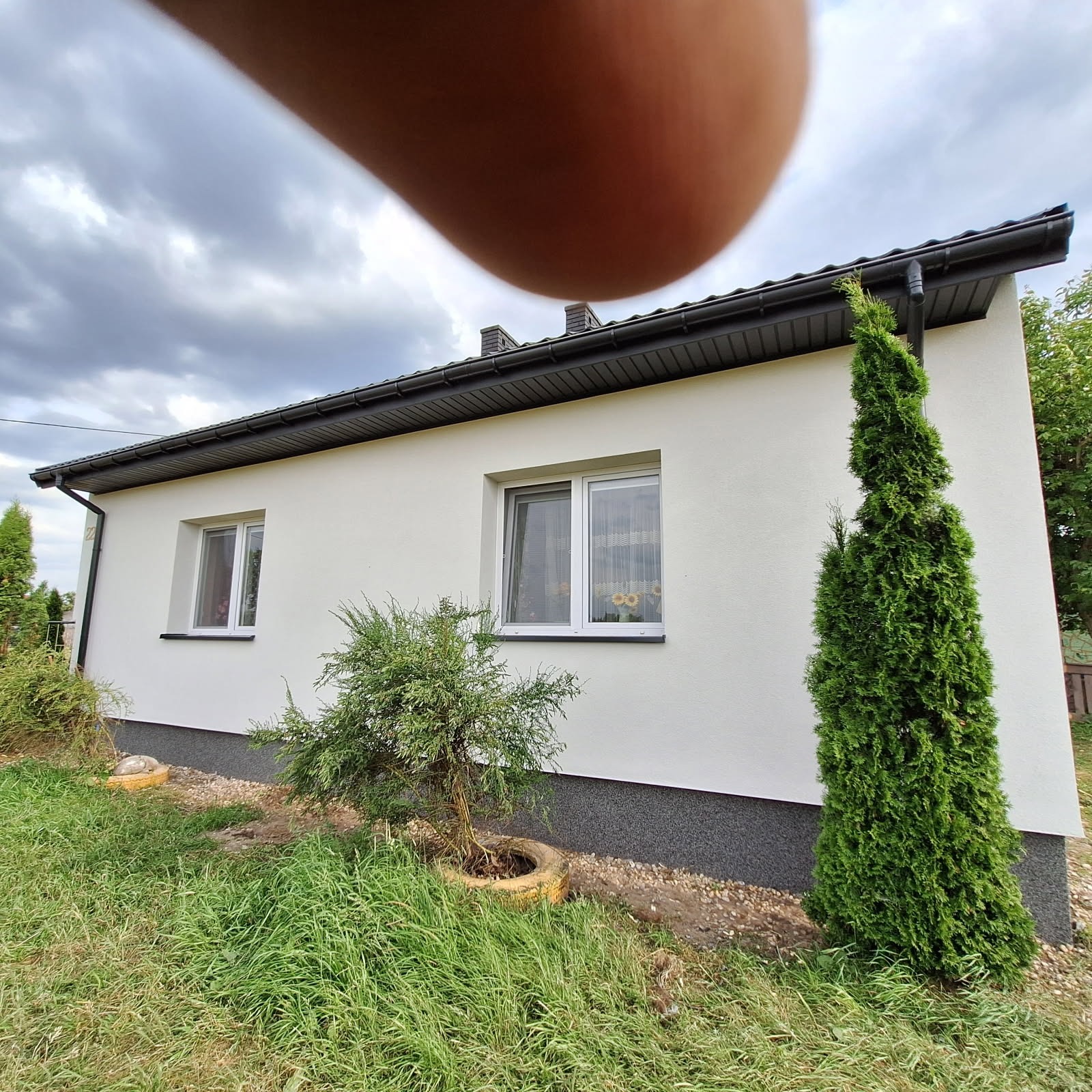 Biała elewacja domu jednorodzinnego z szarą podmurówką, widoczna zieleń trawnika i krzewów. U góry kadru widoczny fragment palca.