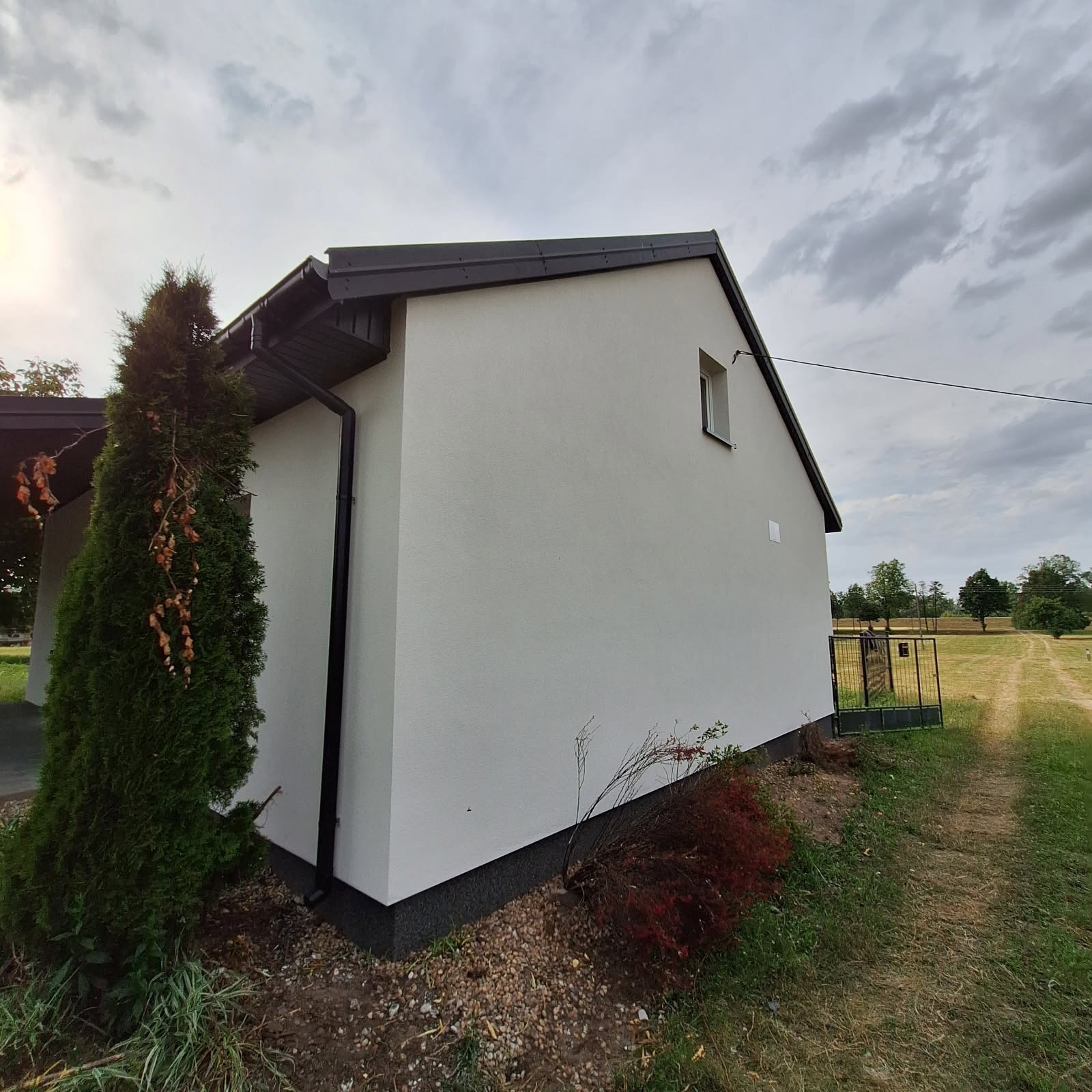 Jasnoszara elewacja domu jednorodzinnego z czarnym dachem i rynnami, widok z boku, na pierwszym planie iglak, w tle brama wjazdowa i polna droga.