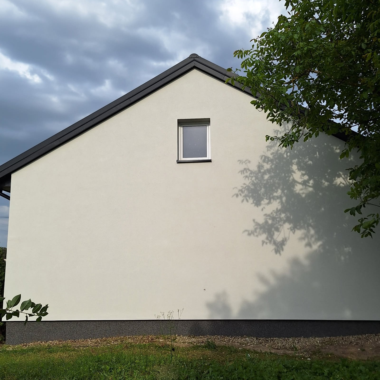 Ściana domu z oknem, po tynkowaniu, z fragmentem dachu i cieniem drzewa. Jasna elewacja kontrastuje z ciemnym cokołem i dachówką.
